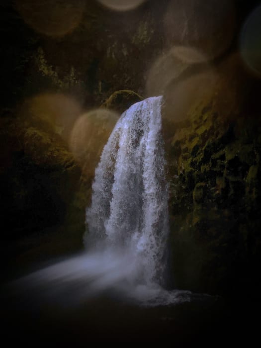 Scenic waterfall cascading down mossy rocks into a dark pool, captured with a soft focus and bokeh in an outdoor setting.