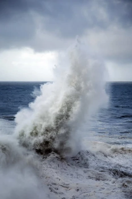 Dramatic ocean wave crashing and spraying water in an outdoor, overcast seascape environment with a dark blue sea and cloudy sky.