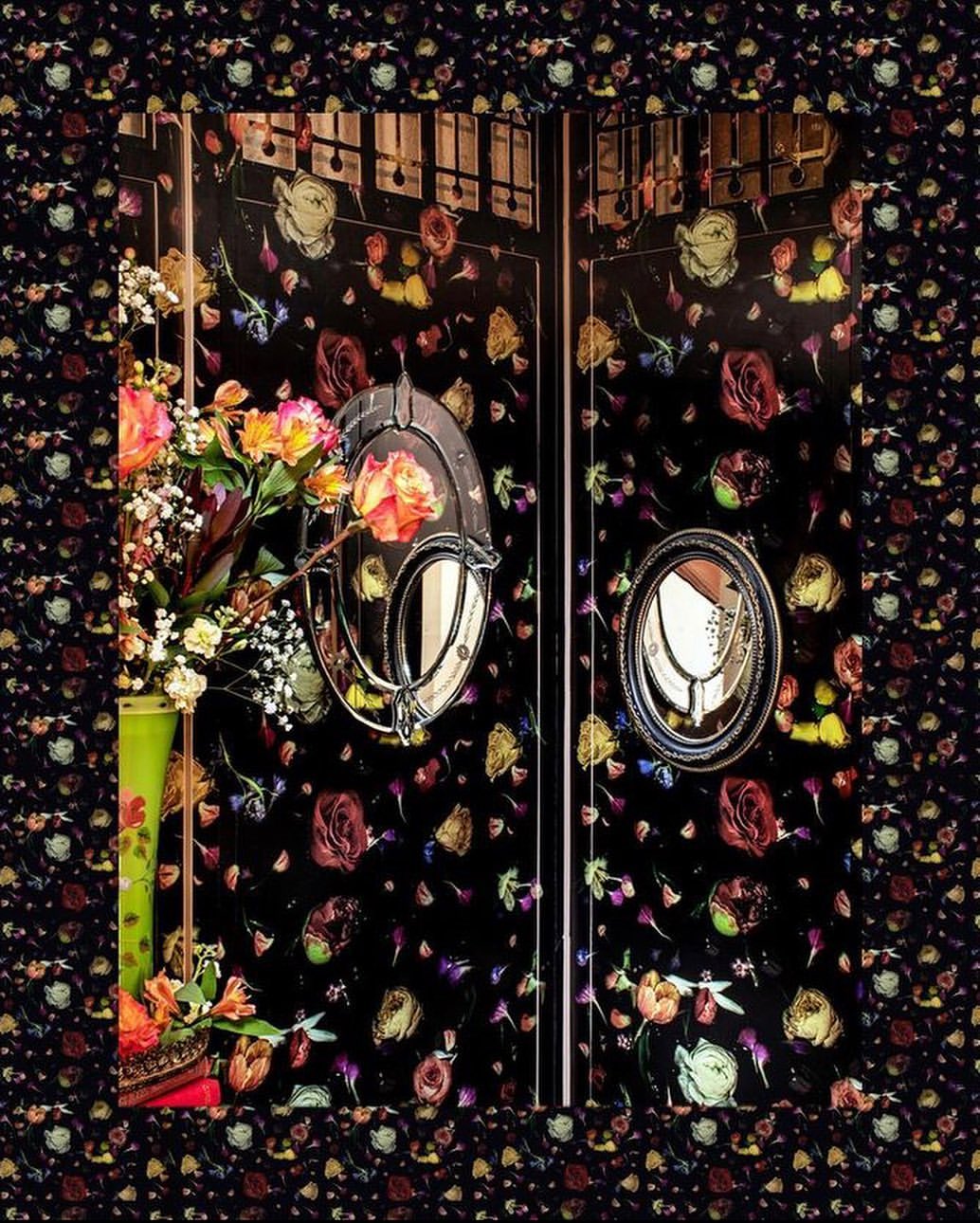 Ornate floral arrangement in a tall vase sits before a dark floral patterned wall with vintage mirrors adding depth and elegance.