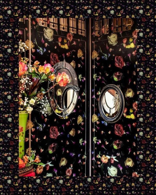 Ornate floral arrangement in a tall vase sits before a dark floral patterned wall with vintage mirrors adding depth and elegance.
