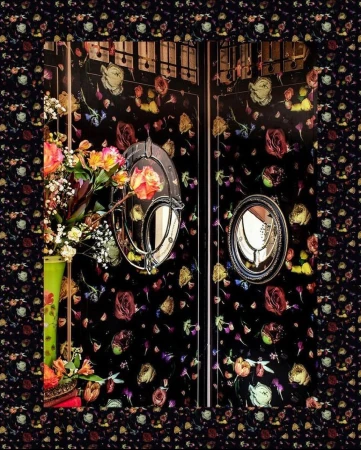 Ornate floral arrangement in a tall vase sits before a dark floral patterned wall with vintage mirrors adding depth and elegance.