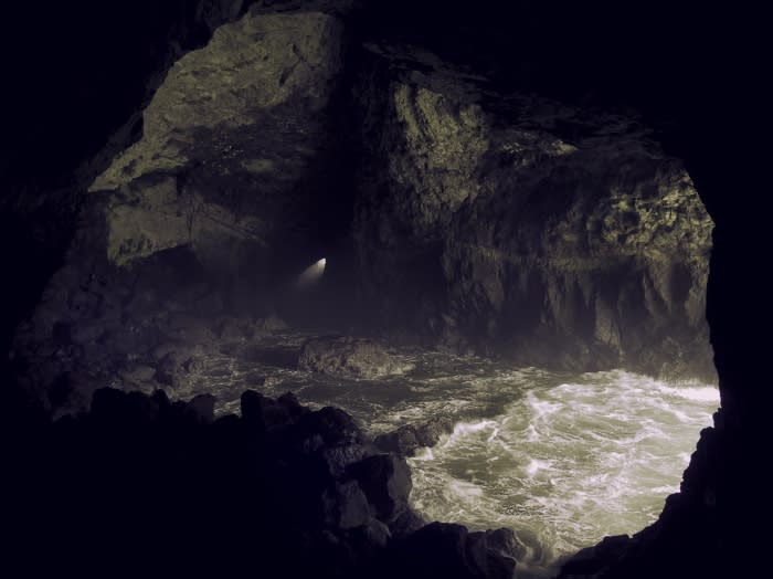 Cave opening with ocean waves crashing against rocks inside, illuminated by a light source in the distance, creating a dramatic scene.