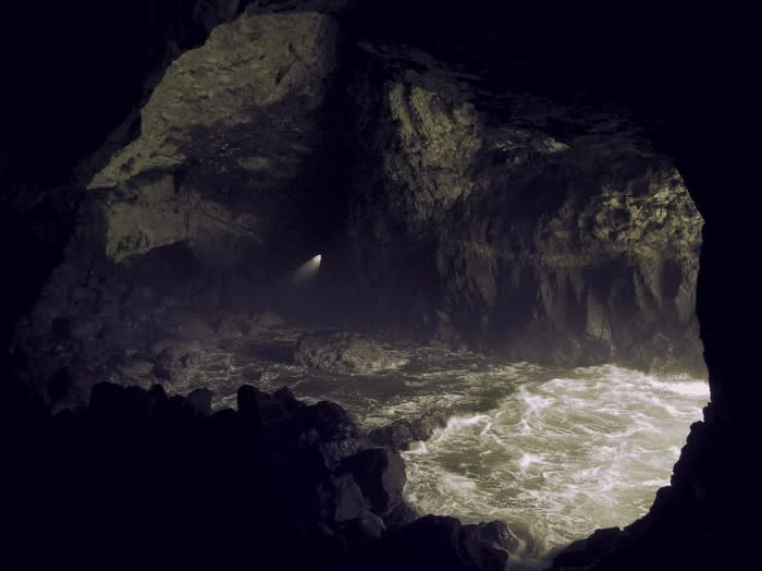 Cave opening with ocean waves crashing against rocks inside, illuminated by a light source in the distance, creating a dramatic scene.