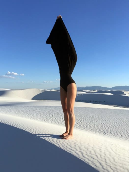 Person in a black bodysuit pulling a dark fabric over their head standing on white sand dunes under blue sky