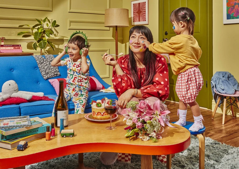 Smiling mother with face stickers sits at a coffee table with two children in a brightly lit living room with playful decor.