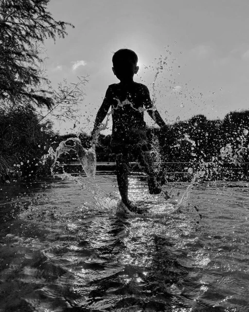 Black and white silhouette of a child running through shallow water, creating splashes against a sunlit background with silhouetted trees.
