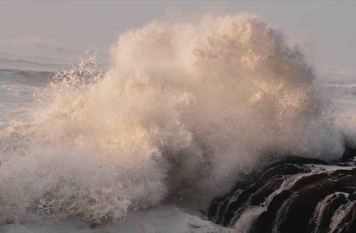 Dramatic seascape featuring a large wave crashing against dark rocks, showcasing the ocean's power and raw beauty in a coastal setting.