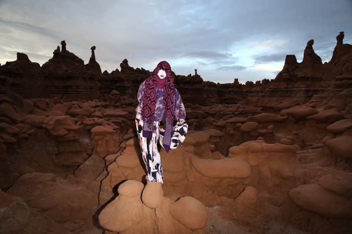 Fashion model wearing hooded outfit poses amid otherworldly rock formations at twilight. Abstract clothing, outdoor, and landscape setting.