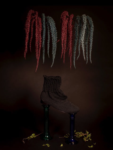 Dark, conceptual still life of a black knitted slipper perched on two colored glass vases, beneath hanging, textured strands on dark background.