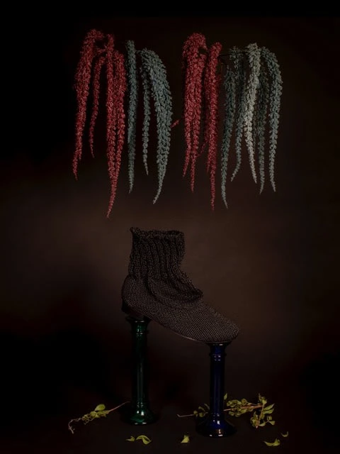 Dark, conceptual still life of a black knitted slipper perched on two colored glass vases, beneath hanging, textured strands on dark background.