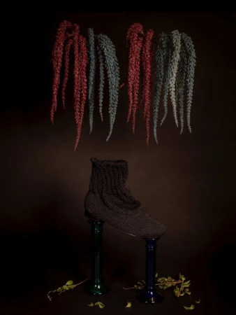 Dark, conceptual still life of a black knitted slipper perched on two colored glass vases, beneath hanging, textured strands on dark background.