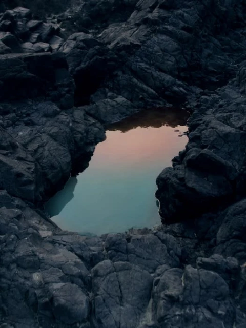 Eroded dark volcanic rock formation frames a tide pool filled with turquoise water reflecting a soft pink and orange sky at dusk.
