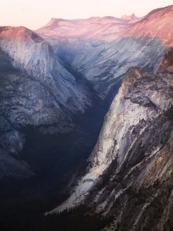 Yosemite Valley landscape view with gray granite rock formations, trees, and pinkish sky during the day in Yosemite National Park, USA.
