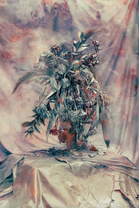 Still life with wilting flowers in a vase on a draped table against a mottled backdrop, soft light, muted colors, artistic.