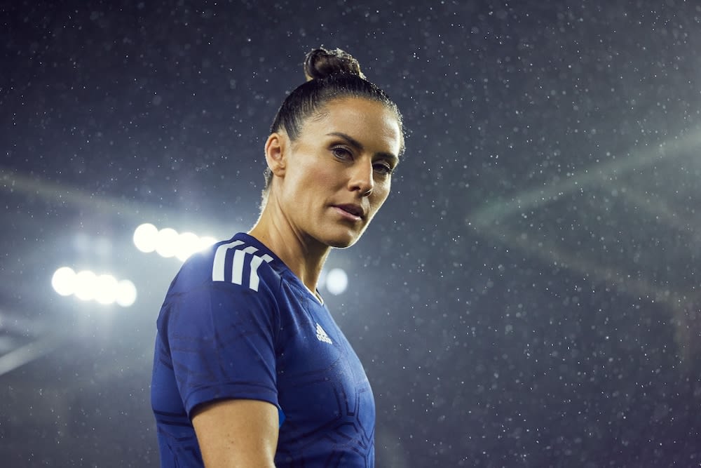 Close-up of athletic adult woman in blue sports top with three stripes, looking towards the camera in a dark, rainy outdoor setting.