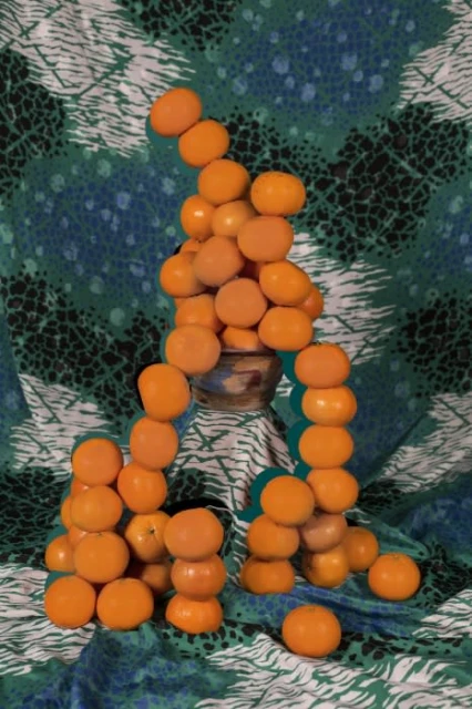 Still life of stacked tangerines next to a vase against a background of blue and green patterned cloth.