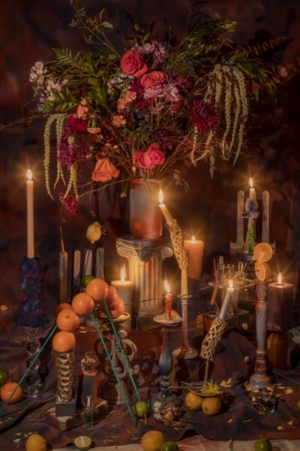 Still life floral arrangement with roses and greenery in a vase surrounded by lit candles and various fruits like lemons and oranges.