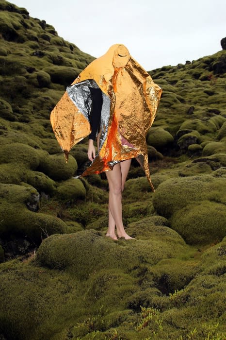 Person stands in a mossy, outdoor landscape, draped in a shiny orange and silver reflective blanket that obscures most of the figure.