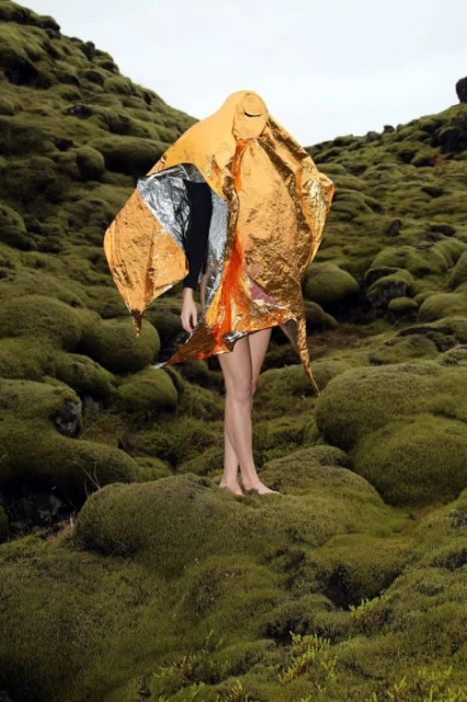 Person stands in a mossy, outdoor landscape, draped in a shiny orange and silver reflective blanket that obscures most of the figure.