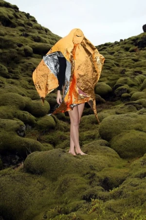 Person stands in a mossy, outdoor landscape, draped in a shiny orange and silver reflective blanket that obscures most of the figure.