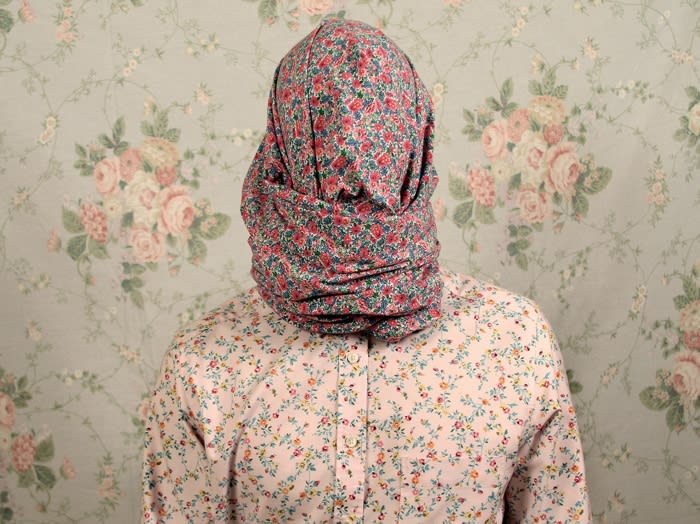 Person covered in floral fabric stands against a matching floral wallpaper in a studio environment with a soft, muted light.