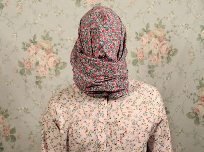 Person covered in floral fabric stands against a matching floral wallpaper in a studio environment with a soft, muted light.
