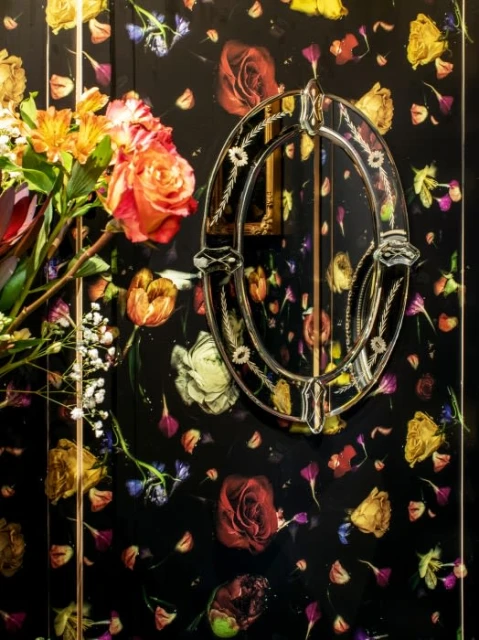 Floral wallpaper with a dark background features an oval mirror with flowers, and a vase of flowers in yellow and orange on the side.