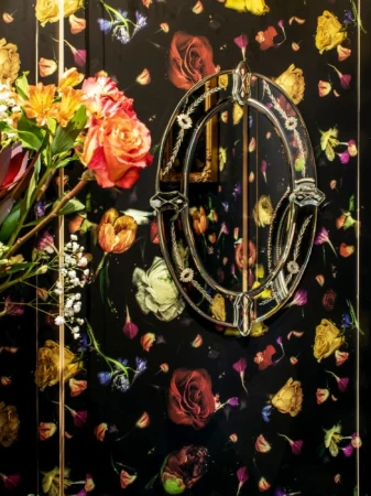 Floral wallpaper with a dark background features an oval mirror with flowers, and a vase of flowers in yellow and orange on the side.