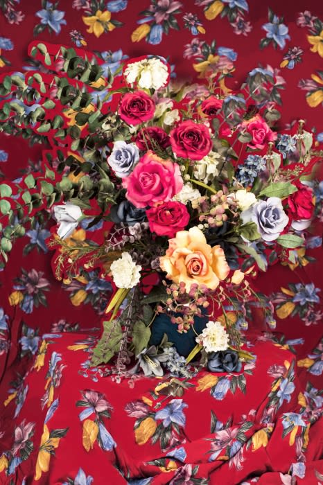 Floral arrangement featuring red, white, and yellow roses with green foliage, presented on a red floral-patterned tablecloth and backdrop.