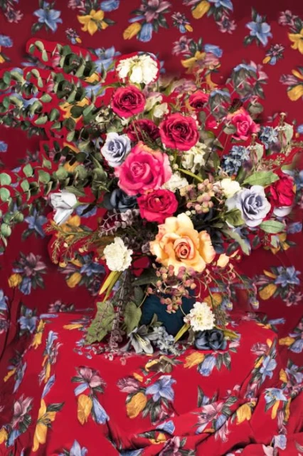 Floral arrangement featuring red, white, and yellow roses with green foliage, presented on a red floral-patterned tablecloth and backdrop.