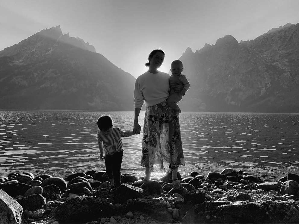 Black and white landscape featuring a mother holding a baby, standing near the water with a toddler, mountains in the background.