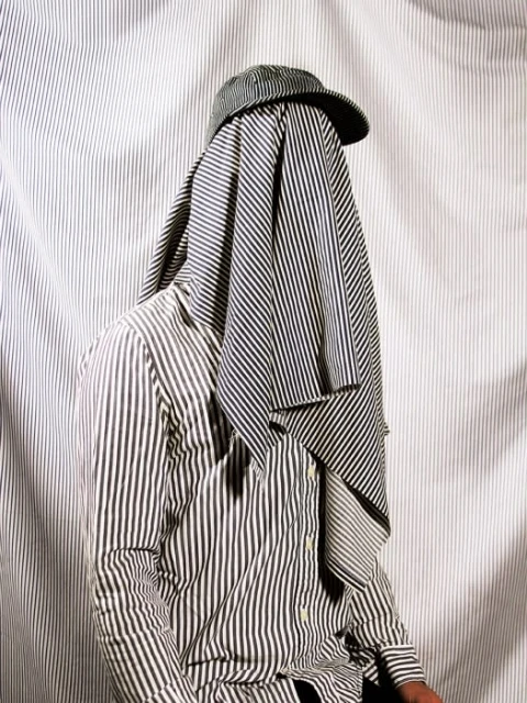 Person draped in a striped fabric and wearing a striped cap stands against a striped background, creating a cohesive monochrome look.