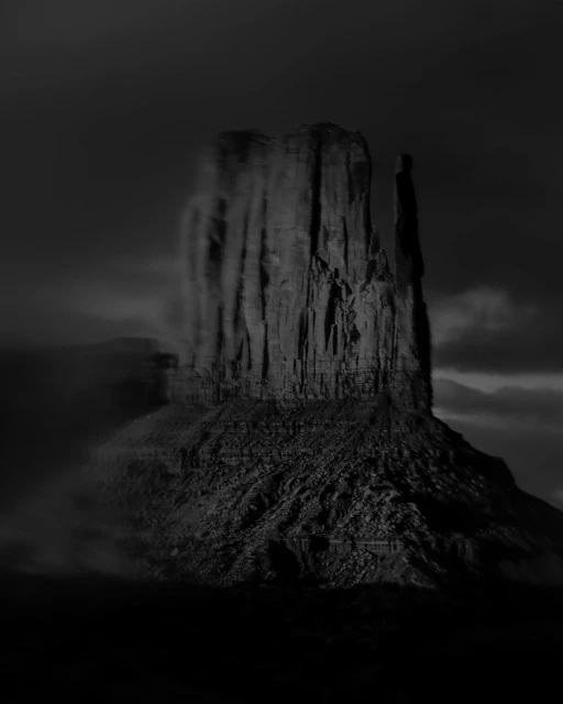 Monochromatic landscape of a large rock formation, possibly a butte or mesa, shrouded in mist and fog under a dark, cloudy sky.