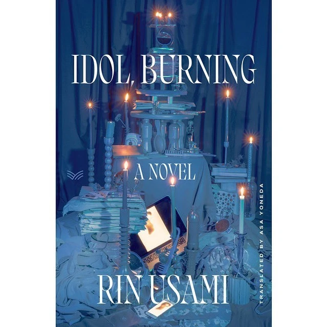 Book cover of 'Idol, Burning' by Rin Usami, featuring a curated collection of objects, including candles, an open laptop, and drapes.