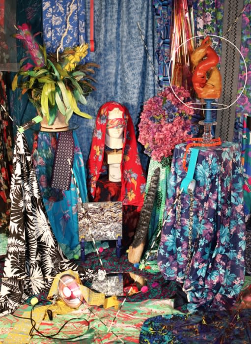 Surreal floral arrangement featuring a mannequin head in a red headscarf surrounded by a tapestry of floral fabrics and sculptures.