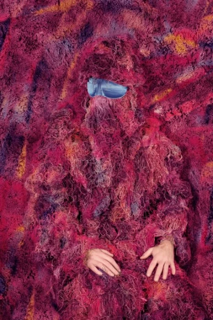 Figure completely covered in a pink, orange, and purple shag-carpet-like material with a pair of silver sunglasses. Hands are visible.