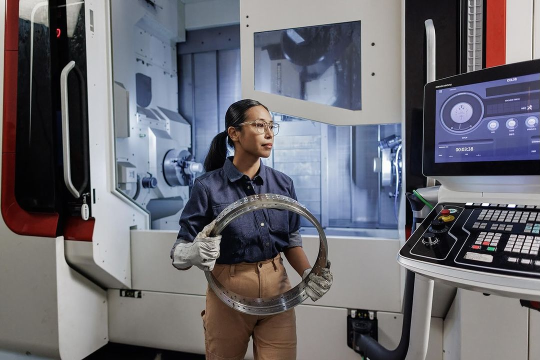 An Asian woman in a factory holds a metal ring, wearing safety glasses, a blue work shirt, and khaki pants in front of machinery.
