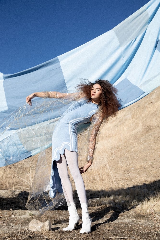Model with curly hair in a light blue dress and white boots posing against a dry hill and large blue fabric banner under a clear sky
