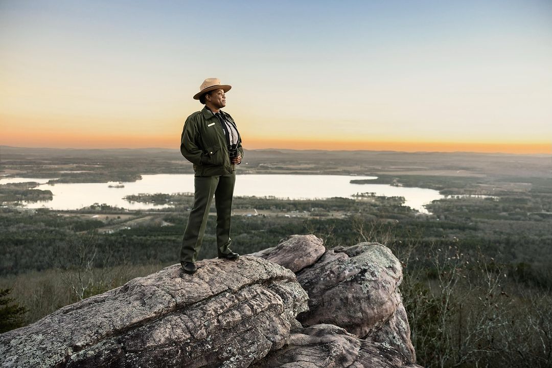 Park ranger stands on a rocky mountain overlooking a scenic landscape with a lake at sunset, wearing uniform and holding binoculars.