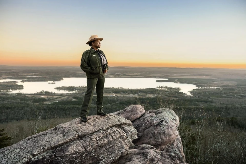 Park ranger stands on a rocky mountain overlooking a scenic landscape with a lake at sunset, wearing uniform and holding binoculars.