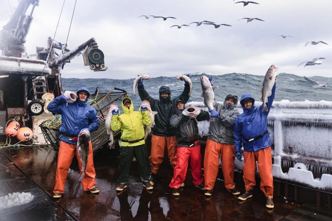 Group of fishermen on a boat deck holding fish, ocean backdrop with birds flying overhead, wearing protective rain gear, overcast day.