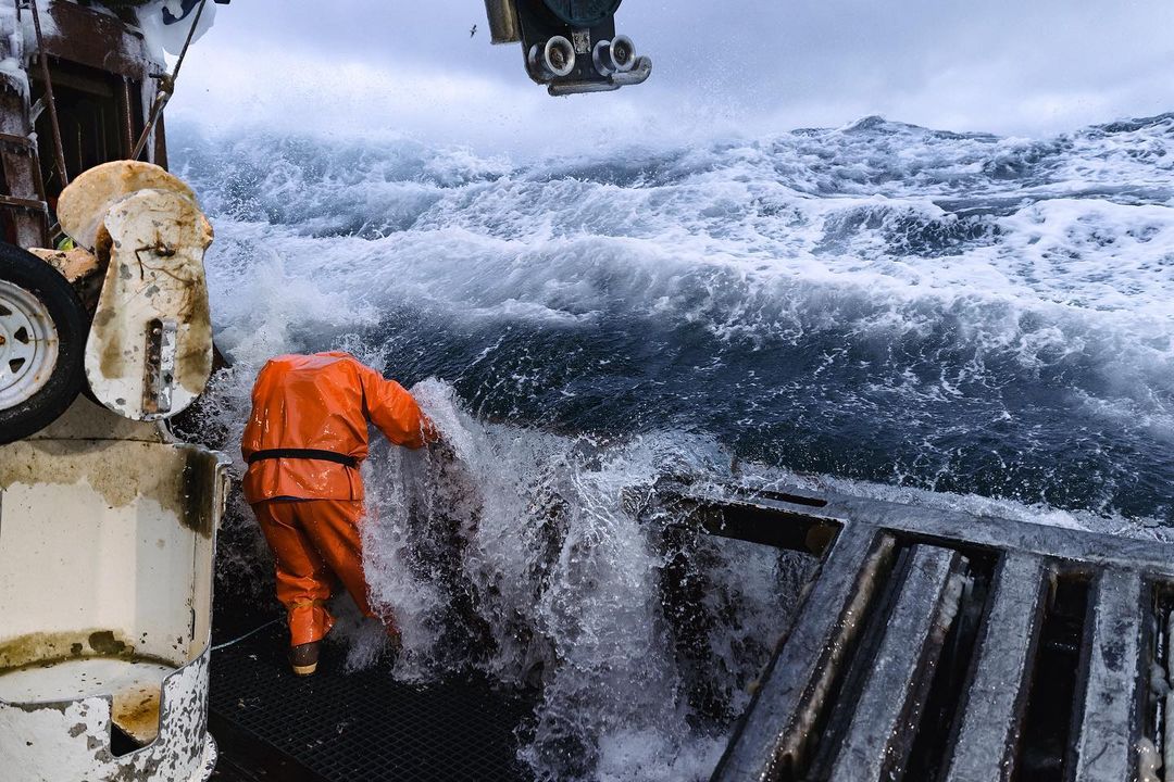 Commercial fisherman in orange waterproofs braves rough ocean waves aboard a fishing boat, showcasing the challenges of the fishing industry.