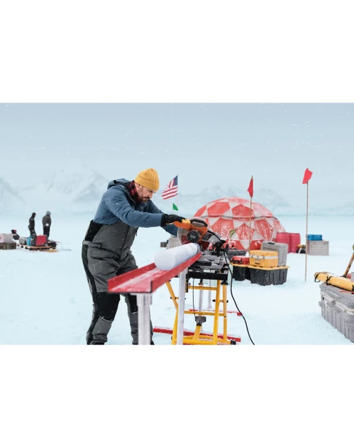 An adult male in snow gear cuts a round object with a power saw in a snowy landscape with a domed building and flags in the background.