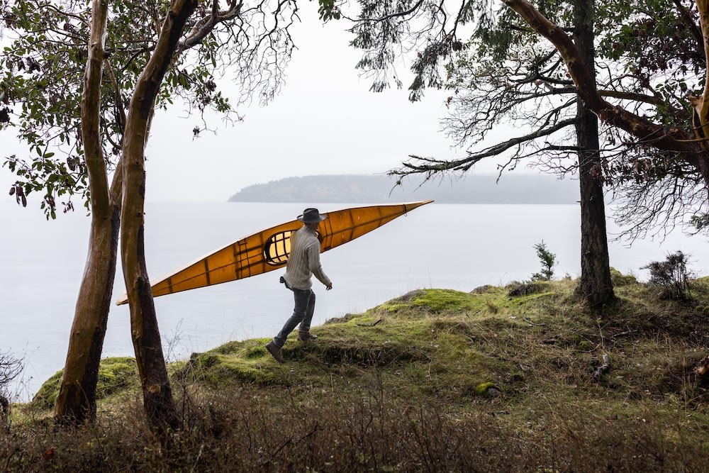 Adult male carrying a yellow kayak over a grassy hill near a body of water, surrounded by trees and cloudy skies in a natural outdoor setting.