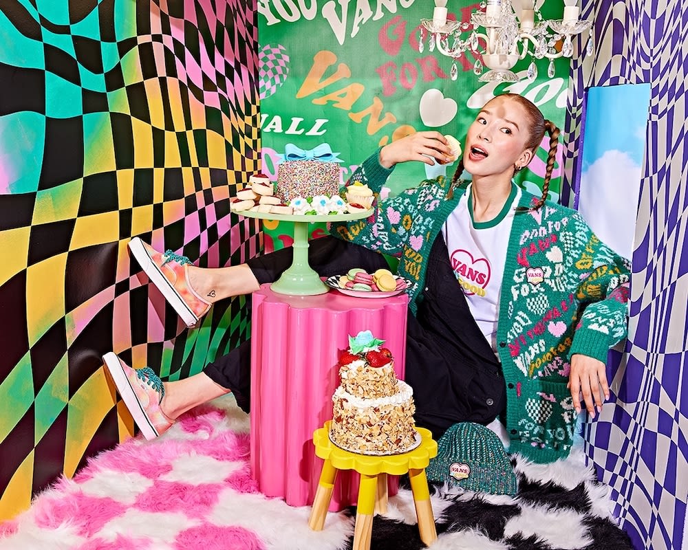An adult female eats pastry in a vivid, patterned photo studio setting featuring Vans branding, cakes, and a fun, youthful aesthetic.