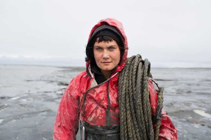 Adult female in red hooded waterproof jacket holds rope on shoulder, standing on wet, muddy tidal flats under an overcast sky.