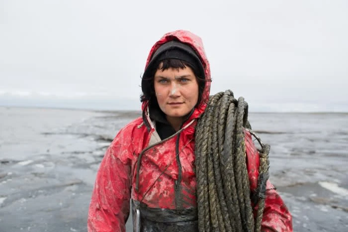 Adult female in red hooded waterproof jacket holds rope on shoulder, standing on wet, muddy tidal flats under an overcast sky.