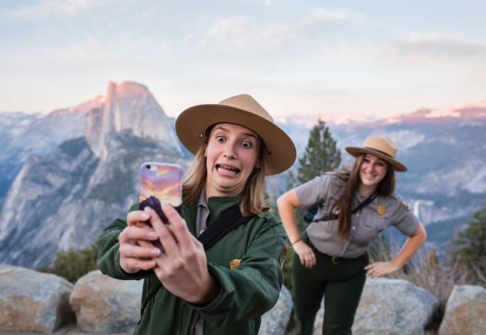 Two park rangers take a selfie in Yosemite National Park with Half Dome mountain in the background on a clear day with sunset lighting.