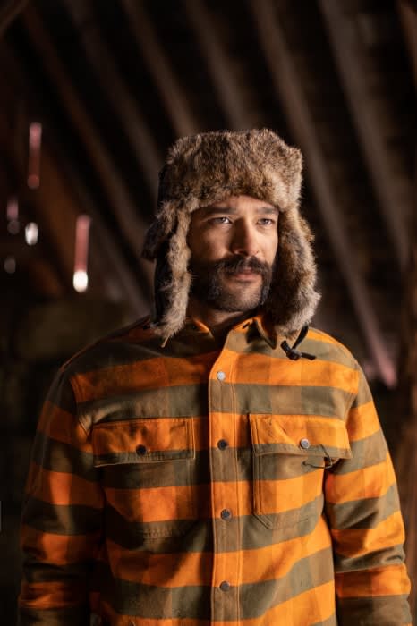 Man in fur hat and striped shirt in rustic interior, looking off-camera. Natural light highlights textures and warm tones.