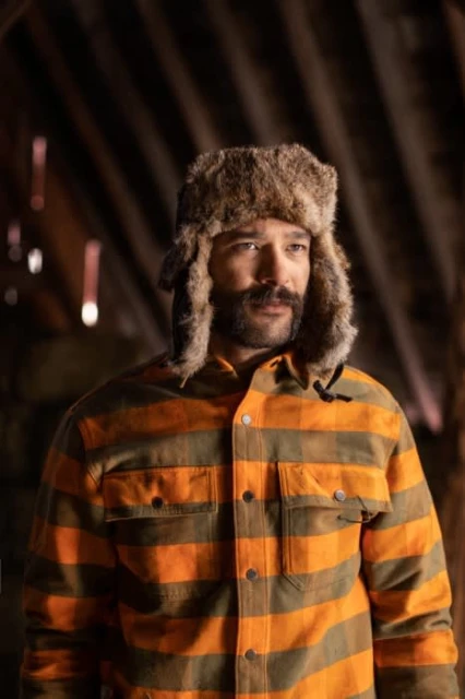 Man in fur hat and striped shirt in rustic interior, looking off-camera. Natural light highlights textures and warm tones.